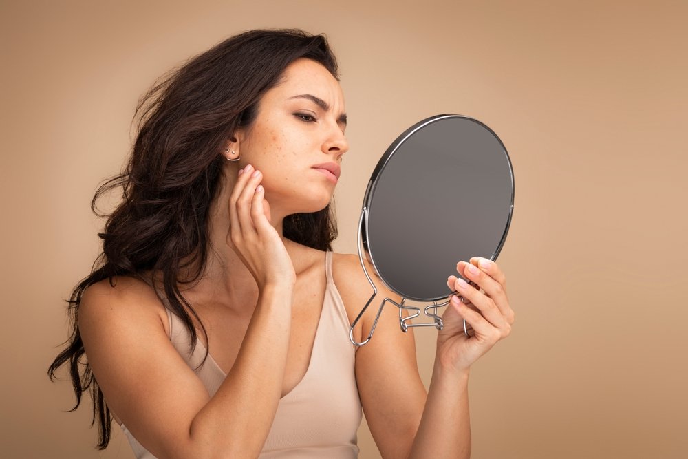 A middle-aged woman looking sad as she inspects her skin in a hand-held mirror.