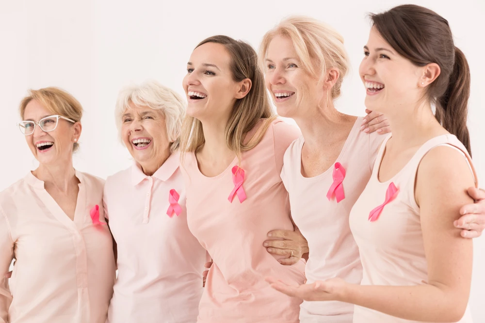 Group of five women of different ages smiling and standing together, each wearing a pink top with a breast cancer awareness ribbon.