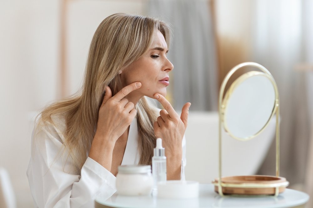 An unhappy blond, middle-aged woman is inspecting problem skin in a mirror.