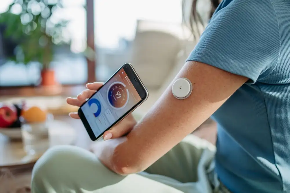 Woman wearing a continuous glucose monitor on her upper arm while checking real-time blood sugar data on a smartphone app in a home setting.