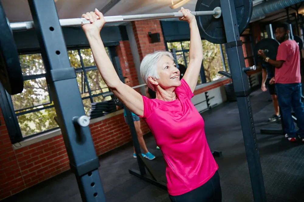 Older woman lifting a barbell overhead during strength training workout in a gym, demonstrating muscle-building exercise for healthy aging and weight loss support